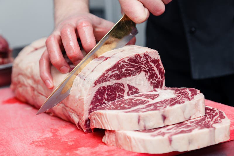 Closeup macro cook`s hand cut ribeye marbled beef steak with knife on red plastic cutting board on metal table in restaurant kitchen. Concept packing semi-finished products, steakhouse, grill meat. Closeup macro cook`s hand cut ribeye marbled beef steak with knife on red plastic cutting board on metal table in restaurant kitchen. Concept packing semi-finished products, steakhouse, grill meat